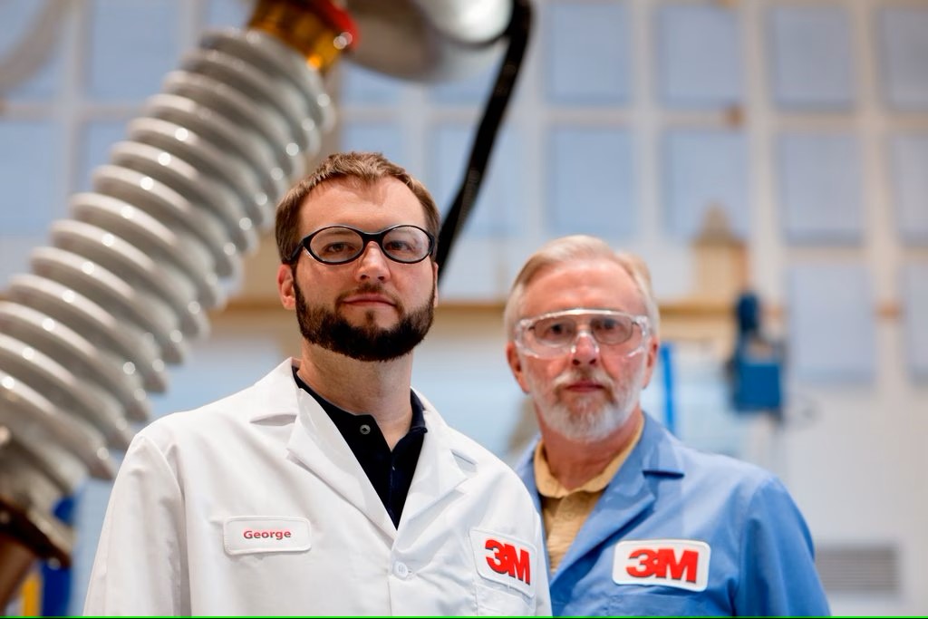 2 men in 3M lab coats standing in front of a transformer 
