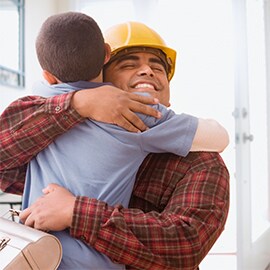 Man wearing hard hat and carrying lunch box smiles and hugs young boy, with open house door in the background