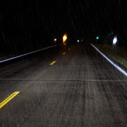 A two-lane highway in the dark and the rain has yellow lines that are brightly illuminated by headlights.