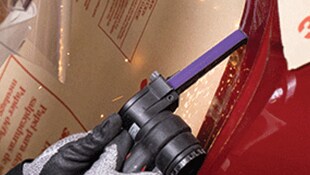 A gloved hand using a handheld power tool with a narrow abrasive belt on a metal surface, producing small sparks
