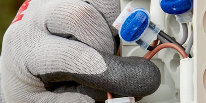 A gloved hand for construction electrical safety holding a blue electrical wire connector inside a white electrical junction box.
