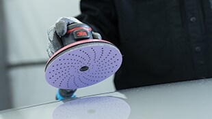 A gloved hand holding a 3M orbital sander with a perforated industrial sanding disc against a smooth surface.
