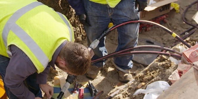 Image of two construction workers handling thick cables at a muddy outdoor worksite.
