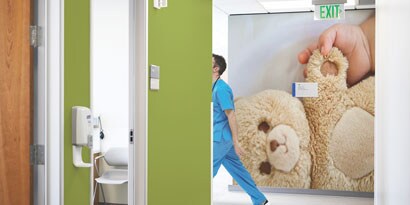 A medical worker strides past a large, inviting wall wrap of a teddy bear held by a baby’s hand.
