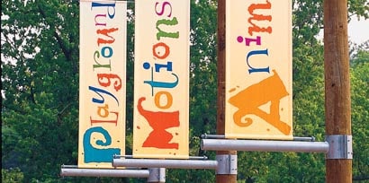 Three wayfinding signs made with 3M grphic films that say playground, motions, and animals on a campground hanging from trees.
