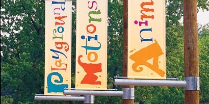 Three wayfinding signs made with 3M graphic films that say playground, motions, and animals on a campground hanging from trees.
